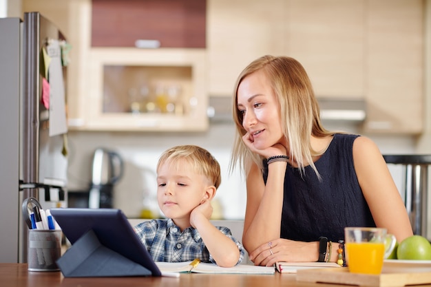 Mutter und Sohn schauen sich Webinare oder Online-Kurse auf dem Tablet-Computer an, wenn sie zu Hause am Küchentisch sitzen