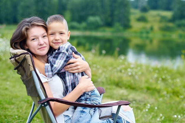 Mutter und Sohn sitzen auf Stuhl und entspannen in der Natur nahe dem See. Outdoor-Aktivitäten im Sommer.