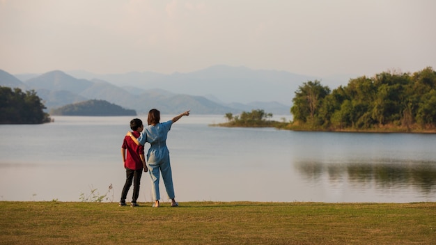 Mutter und Sohn stehen neben großem See und sehen Bergblick im Hintergrund, Mutter zeigt Finger auf Wald. Idee für Familientouristen reist gemeinsam zum Outdoor-Trip.