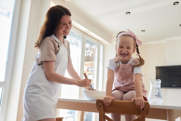 Mutter und Tochter, die einen süßen Kuchen mit Mehlmilch zubereiten, sitzen auf Stühlen an einem Tisch in einem modernen