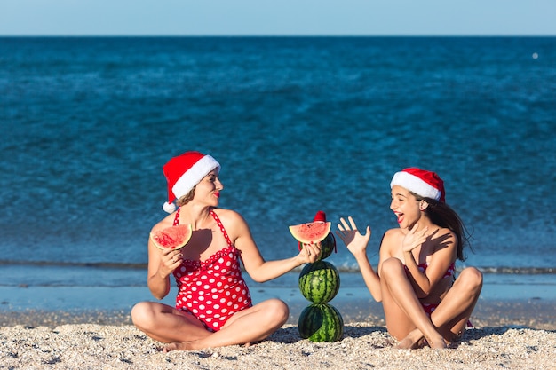 Mutter und Tochter feiern Sommerweihnachten am Strand und essen Wassermelone. Sommerschneemann aus Wassermelonen.