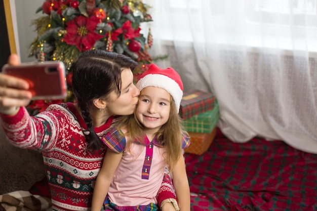 Mutter und Tochter in der Nähe des Weihnachtsbaumes machen Fotos und Selfies am Telefon