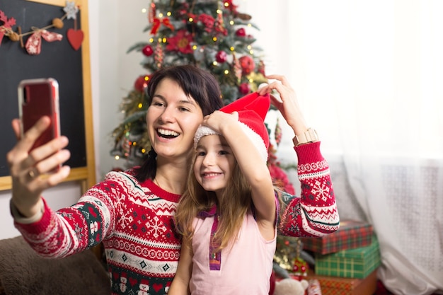 Mutter und tochter in der nähe des weihnachtsbaums machen fotos und selfies am telefon und kommunizieren per video