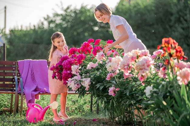 Mutter und Tochter in einem leinenrosa Kleid kümmern sich um Pfingstrosen im Garten, gießen die Blumen. Er lächelt perfekt und lacht. Beleuchtet von den Strahlen der untergehenden Sonne am Abend.