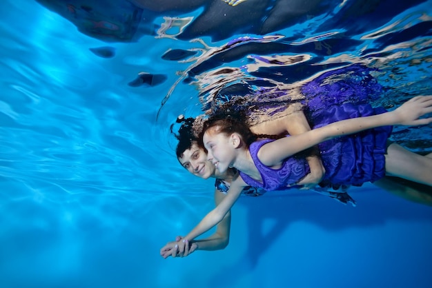 Mutter und Tochter in lila Kleid schwimmen vorwärts und umarmen sich unter Wasser im Pool auf blauem Hintergrund