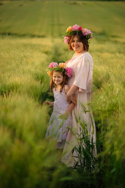 Mutter und Tochter in rosa Kleidern in einem grünen Weizenfeld. Auf ihren Köpfen haben sie Blumenkränze