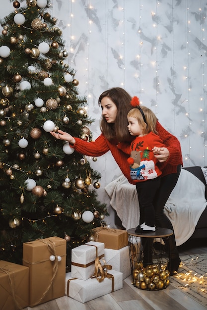 Mutter und Tochter in roten warmen Pullovern schmücken den Weihnachtsbaum. Glückliche Mutterschaft. Warme familiäre Beziehungen. Weihnachten und Silvester Interieur. Liebe. Familienkonzept.