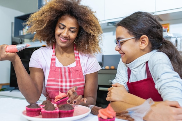 Mutter und Tochter kochen Gebäck in der Küche, lateinamerikanische Erwachsene dekorieren Cupcakes mit Sac a Poche, gefüllt mit rosa Süßigkeitscreme, echter Moment der elterlichen Chilld-Bindung