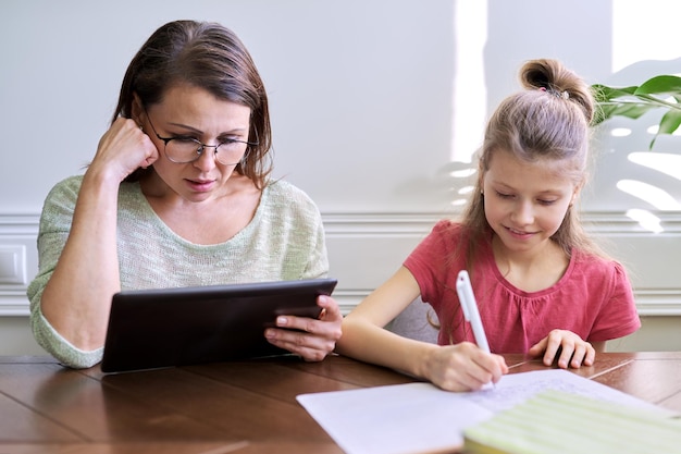 Mutter und Tochter lernen zusammen zu Hause und sitzen am Tisch. Frau mit digitaler Tablette, Internet verwendend, Mädchenschreiben in Notizbuch. Fernunterricht, Eltern helfen Kind Grundschüler