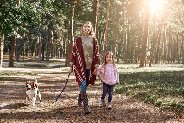 Mutter und Tochter mit Hund wandern im Kiefernwald. Frau trägt einen stylischen Strickmantel, Mädchen trägt einen rosa leuchtenden Pullover