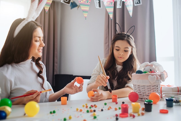 Mutter und Tochter sitzen am Tisch und bereiten sich auf Ostern vor. Sie malen Eier. Mädchen erreichen Farbe mit Pinsel. Junge Frau halten orange Ei.