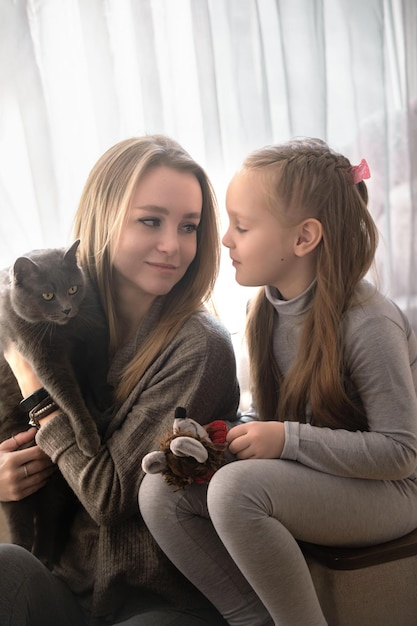 mutter und tochter sitzen zu hause auf der couch, sie haben eine schöne graue katze im arm