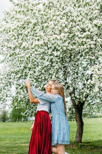 Foto mutter und tochter stehen im frühling oder sommer umarmt unter den blühenden bäumen, roter rock, leichte kleider