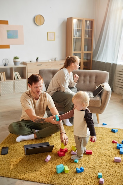 Mutter und Vater benutzen ihre Computer und arbeiten zu Hause mit ihrem Kind, das mit Spielzeug spielt