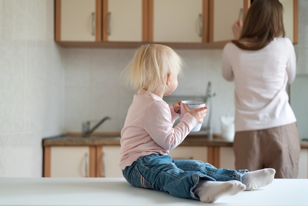 Mutter und zweijähriges Baby mit Tasse in den Händen in der Küche. Familienfrühstück