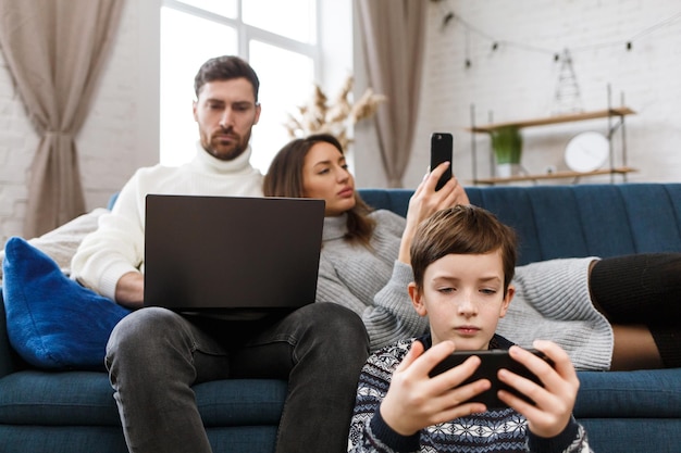 Mutter, Vater und Sohn mit Laptop und Mobiltelefonen zu Hause. Familienmitglieder ignorieren sich gegenseitig und sprechen live. Gadget-Einfluss auf Familienbeziehungen. Moderne Technologien und Suchtkonzept.