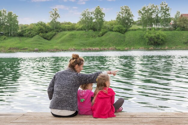 Mutter von zwei Kindern zeigt den Zeigefinger zur Seite auf einem Holzbrett am See sitzend