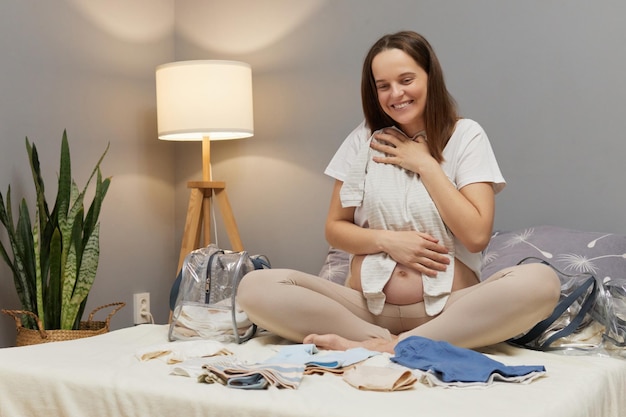 Mutterschaftsmode-Essentials für werdende Mütter Zufriedenheit Charmante regierende Frau bereitet Babykleidung für die Mutterschaftsklinik im Innenraum vor, umarmt den winzigen Schläfer mit Liebe und sanftem Lächeln
