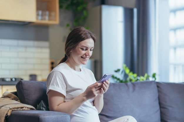 Mutterschaftsurlaub Schwangere, schöne Frau, die zu Hause auf der Couch sitzt und ein Mobiltelefon hält, das Nachrichten eingibt, die anrufen, beobachten, gute Nachrichten empfangen, Lächelnd glücklich