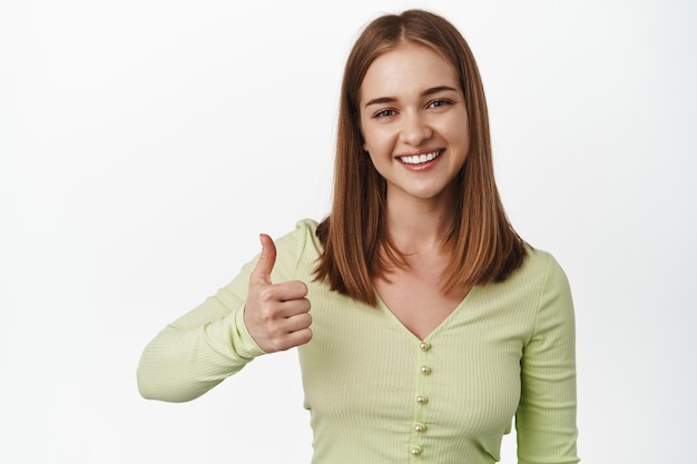 Muy bien. Una joven sonriente muestra el pulgar hacia arriba en aprobación, garantiza la calidad, recomienda un lugar o producto, le gusta y está de acuerdo, comentarios positivos, de pie contra un fondo blanco.