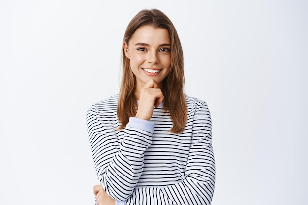 Muy bien. Mujer joven pensativa asiente con la cabeza en aprobación y sonriendo, luciendo determinada y satisfecha, de pie pensativa sobre blanco