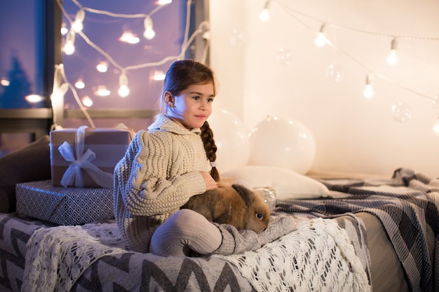 Muy hermosa y encantadora niña de cabello oscuro con suéter blanco sostiene un conejo vivo en el interior de la casa. Año nuevo. Navidad. Liebre. Estudio.