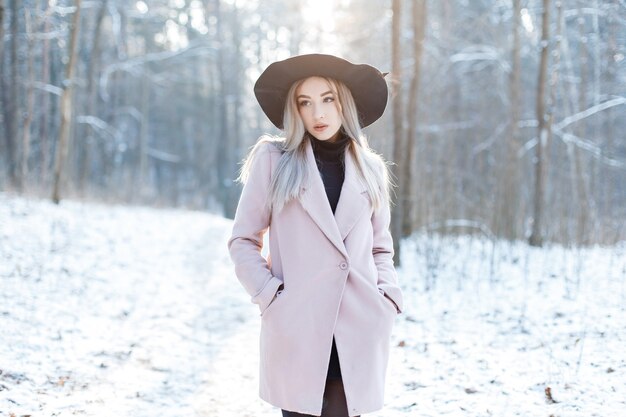 Muy hermosa mujer joven en elegante ropa cálida glamorosa está caminando con un elegante sombrero en un bosque nevado en un día soleado de invierno. Chica moderna atractiva de moda.