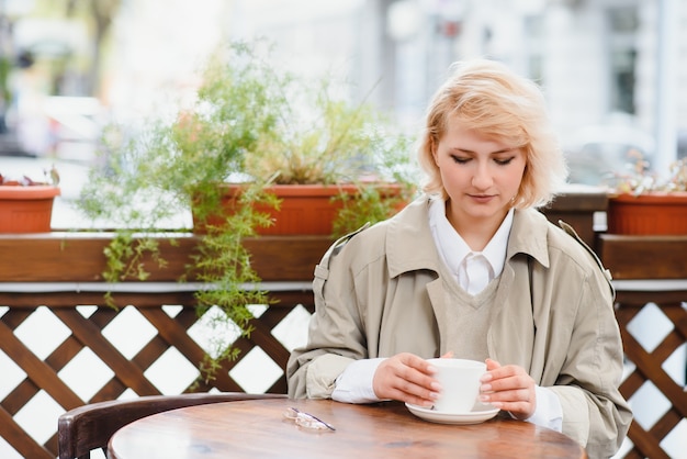 Muy hermosa mujer joven, sentarse en la cafetería y beber café o té, vista frontal de la calle