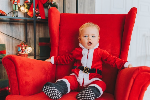 Muy lindo niño pequeño caucásico con traje de Santa en un gran sillón rojo con luces de Navidad