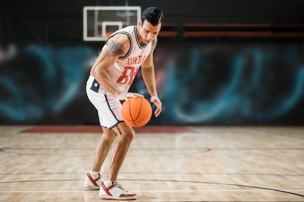 Na Academia. Jovem atlético em uma roupa esportiva branca jogando basquete no ginásio