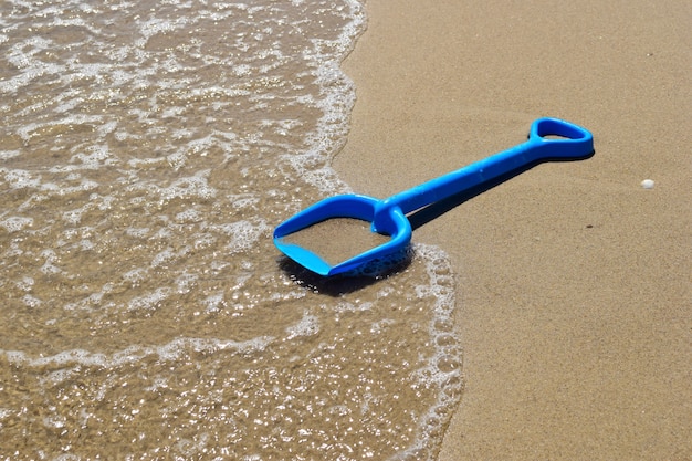 Na areia da praia do mar encontra-se uma pá de criança contra o pano de fundo de uma onda espumosa.