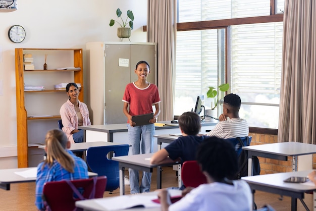 Na escola, um aluno apresenta um projeto aos colegas de classe na sala de aula, uma professora indiana observando