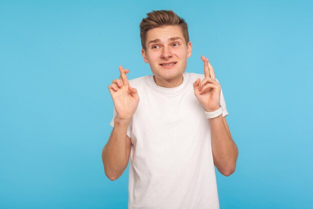 Na expectativa da vitória. retrato de homem de camiseta branca casual cruzando os dedos em gesto de boa sorte e olhando com esperança, desejando sucesso. tiro de estúdio interno isolado em fundo azul