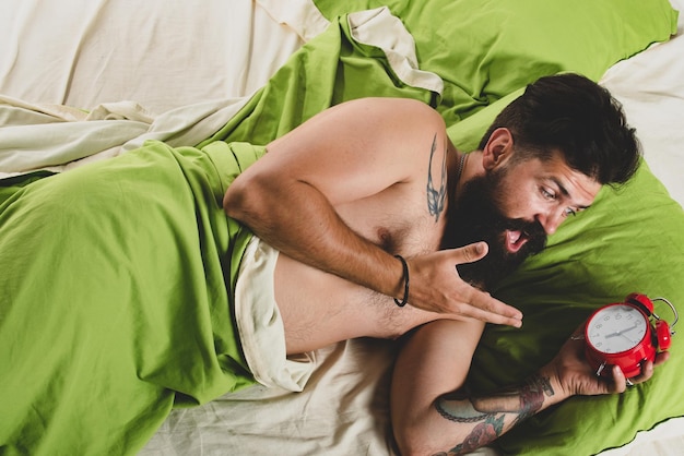 Na hora de acordar, um cara barbudo atraente na cama segurando um alarme nas mãos