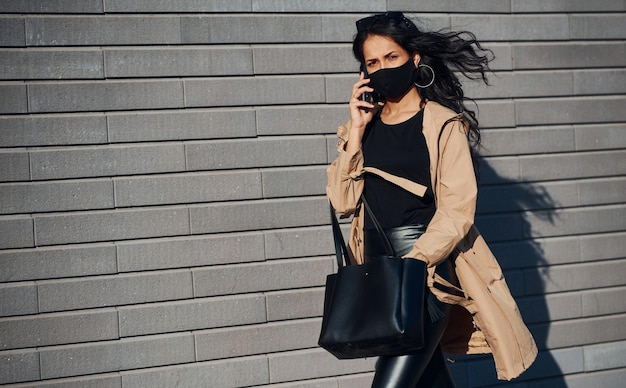 Foto na máscara protetora linda morena com cabelos cacheados e roupas pretas falando por telefone ao ar livre perto da parede