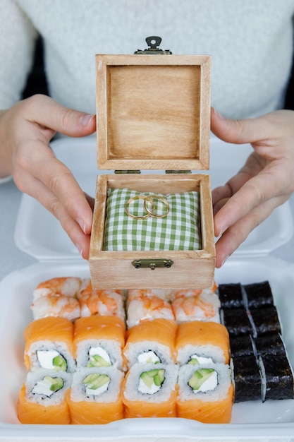 Na mesa da cozinha está uma caixa de rolinhos Filadélfia japoneses Sushi de entrega rápida em um recipiente branco Alianças de casamento nas mãos de uma menina Decoração festiva Conceito de Natal