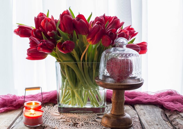 Na mesa em um vaso há muitas tulipas vermelhas, uma vela e um coração