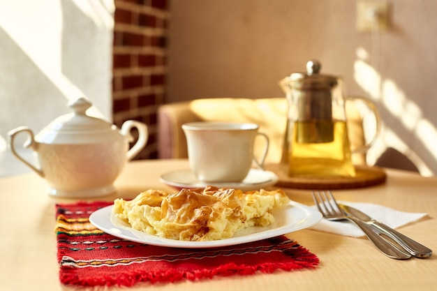 Na mesa está uma omelete, um garfo, uma faca, um bule com chá, um açucareiro, uma chávena e um pires num prato