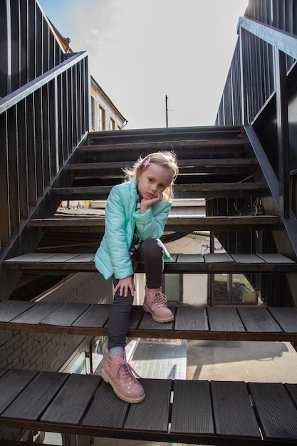 Na moda bonita caucasiana loira menina sentada com um olhar angustiado na escada de ferro, olhando para a câmera.