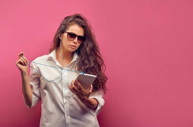 Na moda jovem caucasiana com uma camisa branca em copos com fones de ouvido, segurando um tablet branco nas mãos em uma parede rosa colorida.