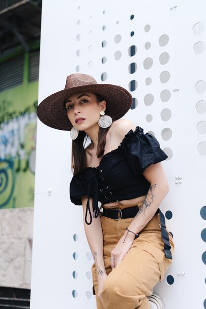 Na moda mulher morena com cabelo comprido, usando um chapéu de vime grande e elegante, posando contra uma parede branca com buracos.