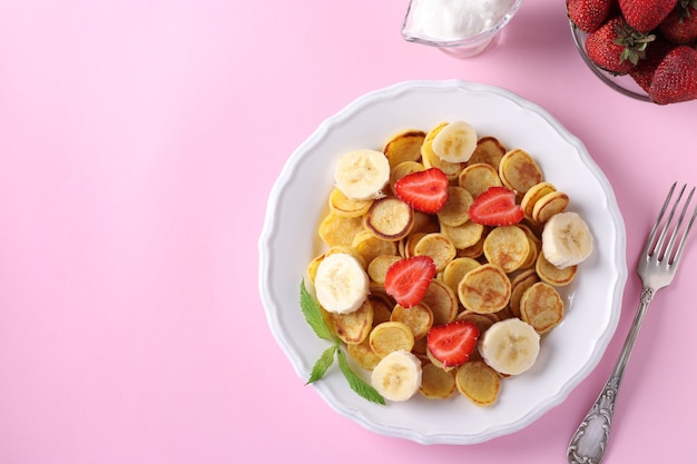 Na moda, pequenas panquecas no café da manhã com morango, banana e creme de leite em chapa branca em fundo rosa. Vista de cima. Copie o espaço para texto ou desenho