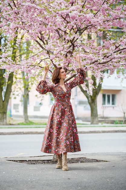 Na primavera, uma mulher caminha por uma rua florida com árvores sakura. Uma garota em um vestido vintage elegante de seda longa caminha entre as árvores floridas
