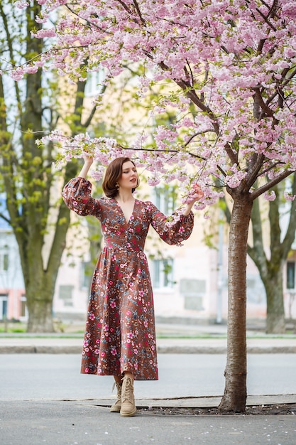 Na primavera, uma mulher caminha por uma rua florida com árvores sakura. Uma garota em um vestido vintage elegante de seda longa caminha entre as árvores floridas