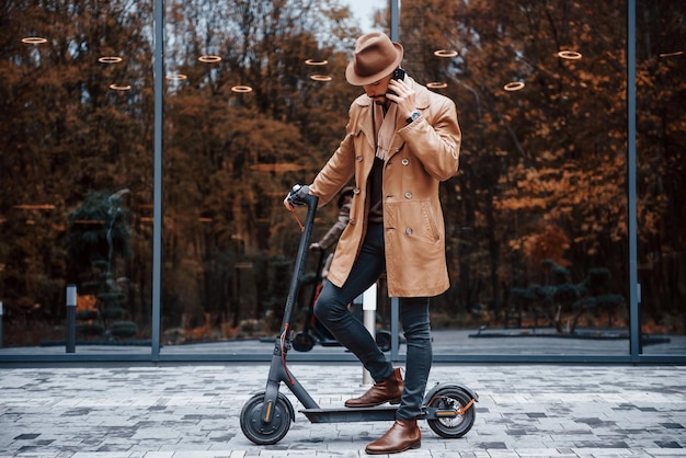 Na scooter elétrica Jovem modelo masculino em roupas da moda está ao ar livre na cidade durante o dia