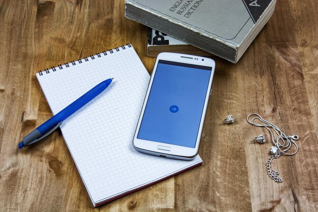 Na superfície de madeira estão livros um caderno com uma caneta um smartphone uma corrente e brincos