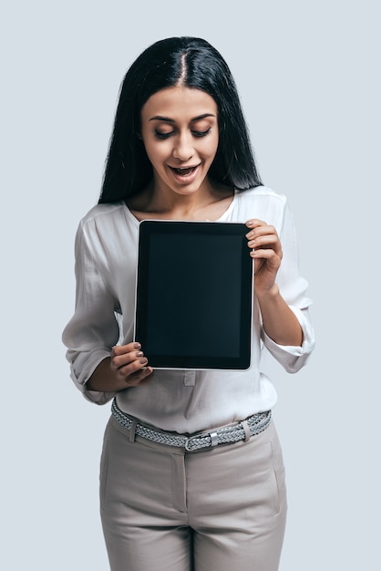 Foto na tendência com tecnologias modernas. mulher jovem e atraente segurando um tablet digital com uma expressão de surpresa em pé contra um fundo cinza