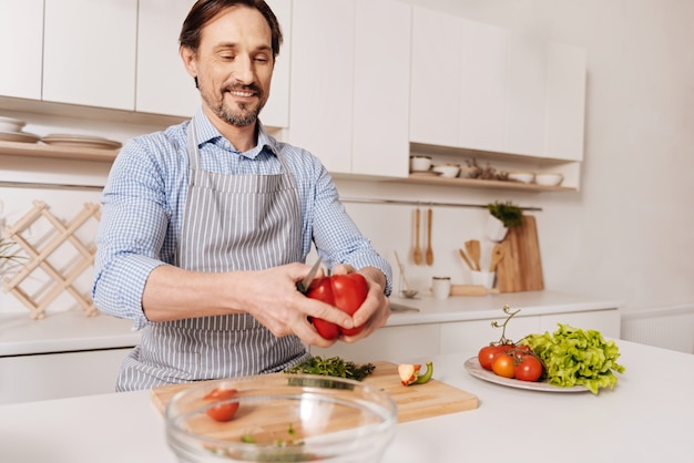 Nach dem Rezept. Kompetenter, erfahrener, fröhlicher Koch, der in der Küche steht und vegetarischen Salat kocht, während er Gemüse schneidet