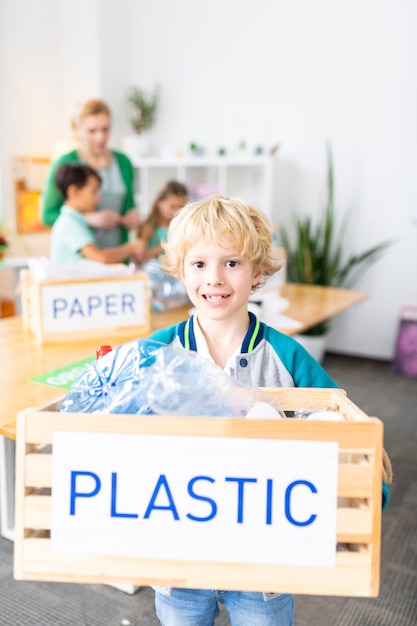 Nach dem Sortieren von Abfällen. Hübscher lustiger Schüler, der eine Kiste mit Plastik hält, nachdem er in der Schule Abfälle sortiert hat