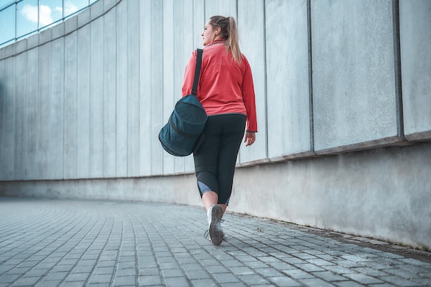 Nach Hause gehen. Rückansicht einer Frau in Übergröße in Sportkleidung, die ihre Tasche trägt und nach Übungen im Freien nach Hause geht. Sportkonzept. Fette Frauen